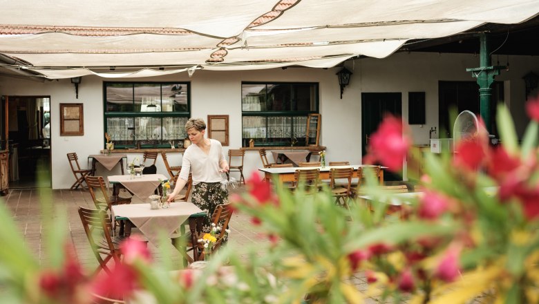 Herbst Inn, © Weinviertel Tourismus / Michael Reidinger A woman prepares tables in a covered outdoor area of an inn. Blurred flowers can be seen in the foreground.
