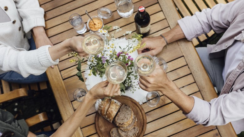 Visit to a wine tavern, © Weinviertel Tourismus GmbH / Sophie Menegaldo Three people clink glasses of wine at a wooden table with bread and flowers on it.