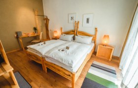 Holzzimmer, © Matthias Auer Ein gemütliches Schlafzimmer mit Holzmöbeln, einem Doppelbett, Nachttischen und einem Schreibtisch. Zwei Lampen und Bilder an der Wand ergänzen das Ambiente.