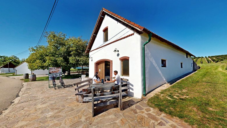 Spritzerrast – Campingstellplatz mit Jause und Panoramablick, © Bernhard Raab Zwei Personen sitzen an einem Holztisch vor einem weißen Gebäude mit rotem Dach, umgeben von grüner Landschaft und Weinbergen.