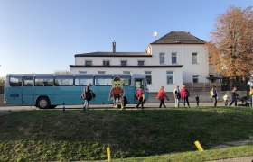 NaturparkBus, © regiobahn RB GmbH Ein blauer Bus steht vor einem Gebäude, Menschen gehen vorbei. Eine bunte Eulenfigur ist im Vordergrund zu sehen.