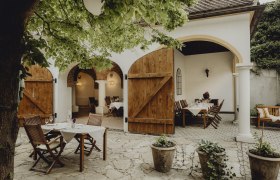 Ein Wirtshaus zum Verweilen, © Niederösterreich Werbung/Sophie Menegaldo Gemütlicher Gastgarten mit Holztischen und Stühlen unter einem Baum, umgeben von weißen Wänden und offenen Holztüren.