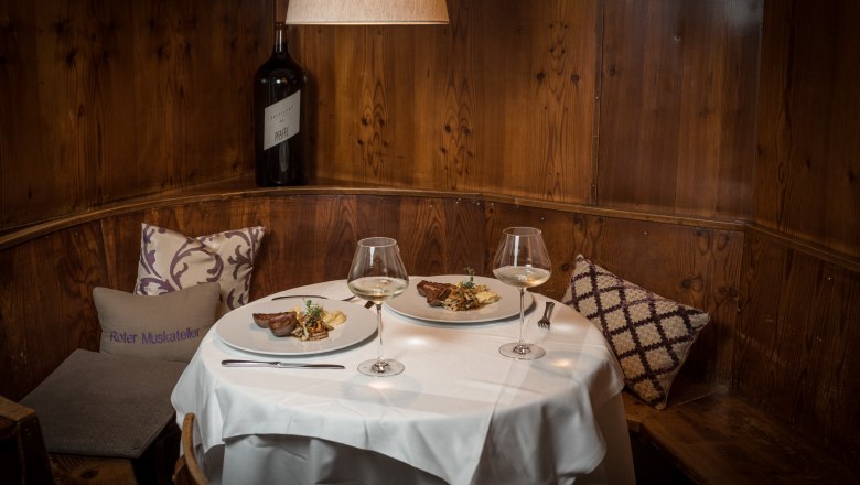 TDR-Hopfeld Dreikönigshof-Gaststube, © Fa. Aumerk Gemütliche Gaststube mit gedecktem Tisch, zwei Tellern mit Speisen und Weingläsern, Holzwand und Kissen.