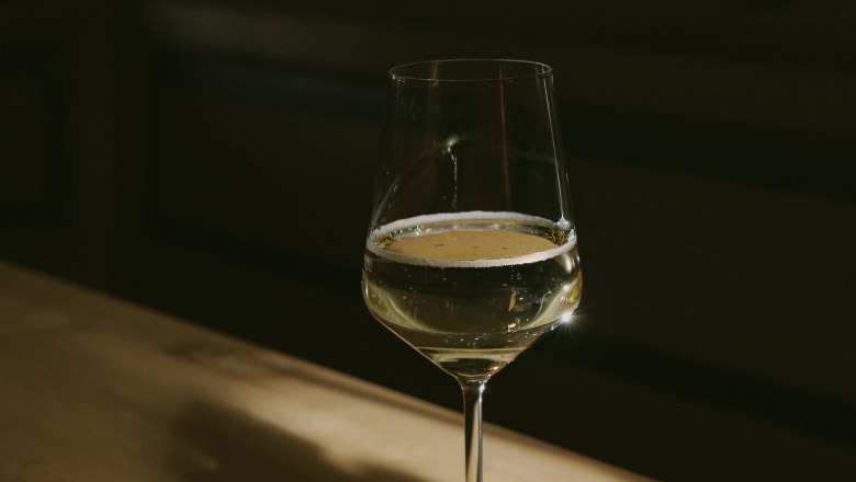 Weinglas, © Weinviertel Tourismus / Sophie Menegaldo Ein Weinglas mit Weißwein steht auf einem Holztisch, beleuchtet von einem warmen Lichtstrahl.