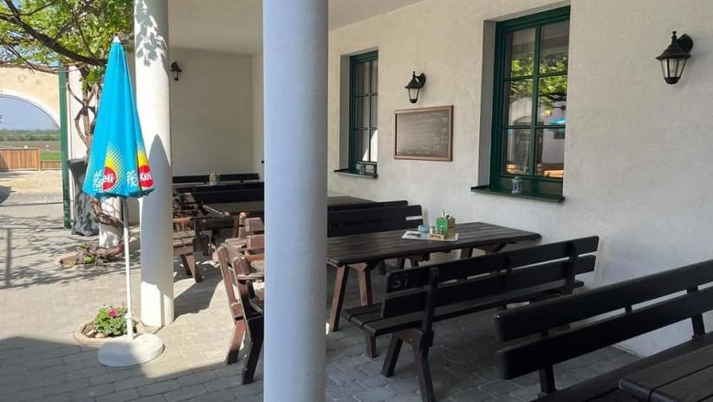 Gastgarten, © Hanfwirt Ein leerer Gastgarten mit Holzbänken und Tischen, einem Sonnenschirm und einer weißen Wand mit Fenstern.