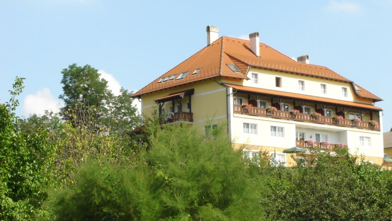 Pension Rudi, © Pension Rudi Ein großes, mehrstöckiges Gebäude mit rotem Dach und Balkonen, umgeben von Bäumen und Pflanzen.