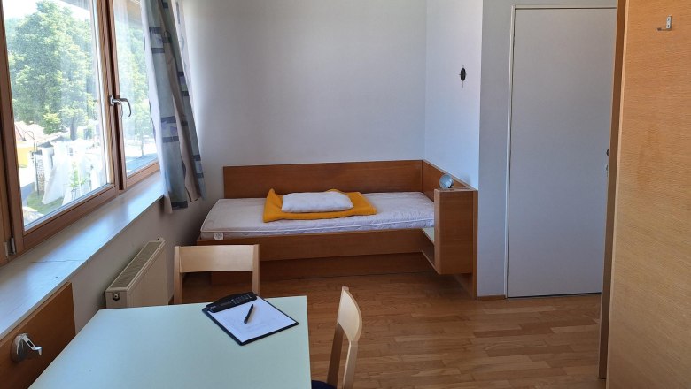 Einzelzimmer, © smart concept team gmbh Ein einfach eingerichtetes Einzelzimmer mit Bett, Tisch und Stuhl, helle Holzmöbel und ein großes Fenster mit Blick nach draußen.