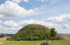 Tumulus im Sommer, © LEADER-Region Weinviertel / Lahofer Ein grasbewachsener Hügel mit einem Kreuz auf der Spitze unter einem bewölkten Himmel.