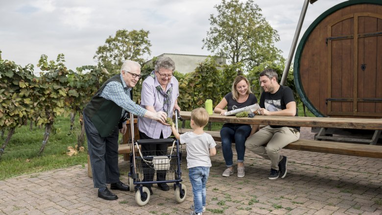 Hohenruppersdorf adventure vineyard, © Weinviertel Tourismus GmbH / Herbst A family sits at a wooden table outside, surrounded by vines. An elderly couple is giving grapes to a child.