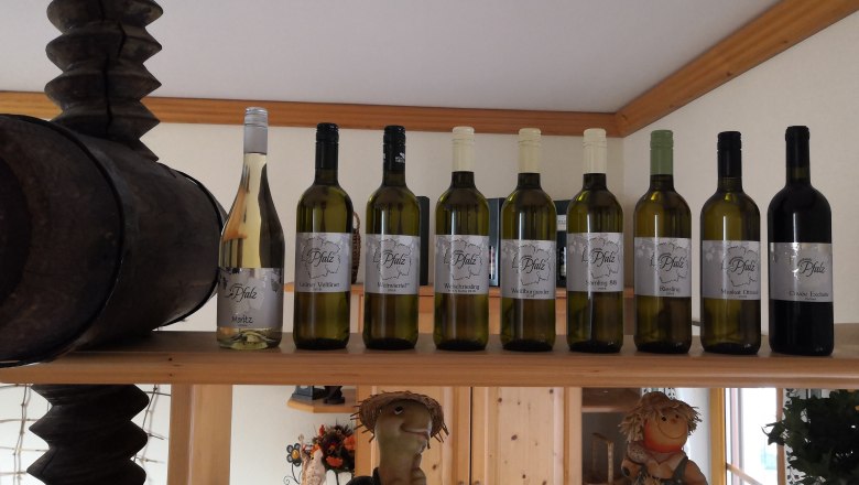 Winzerfamilie Pfalz, © Winzerfamilie Pfalz Weinflaschen auf einem Regal mit Holzfass und Dekofiguren.