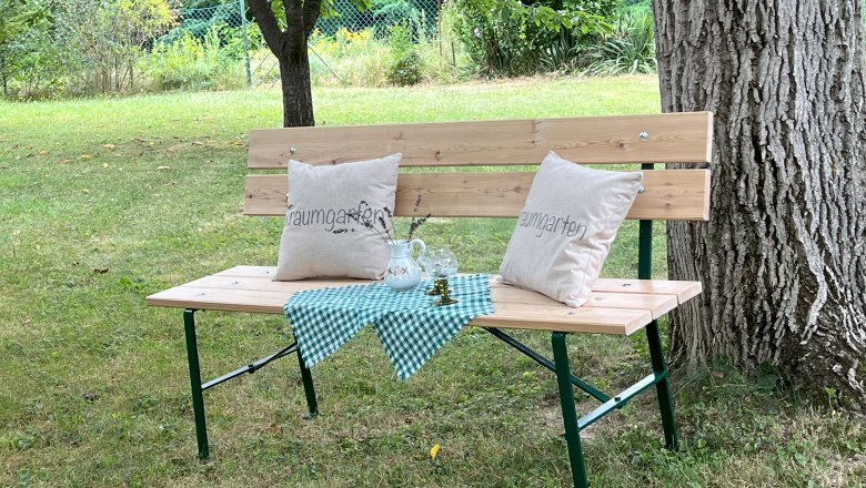 Relaxing under the walnut tree, © Waltraud Forstner Wooden bench with cushions and tablecloth under a tree in the garden.