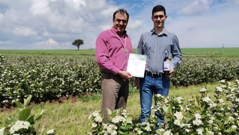 Aroniahof Gepp, © Matthias Gepp Two men stand in a chokeberry field, one holding a certificate, the other a bottle. A tree and blue sky with clouds in the background.