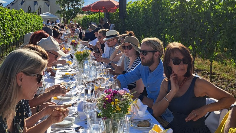 Tafeln im Weinviertel, © Weinviertel Tourismus GmbH Menschen sitzen an einer langen Tafel im Freien neben einem Weinberg, mit einer Kirche im Hintergrund und strahlendem Sonnenschein.