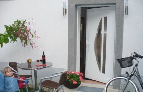 Hereinspaziert, © Anton Dungl Ein offener Hauseingang mit Tisch, Stühlen, Weinflasche und Fahrrad.