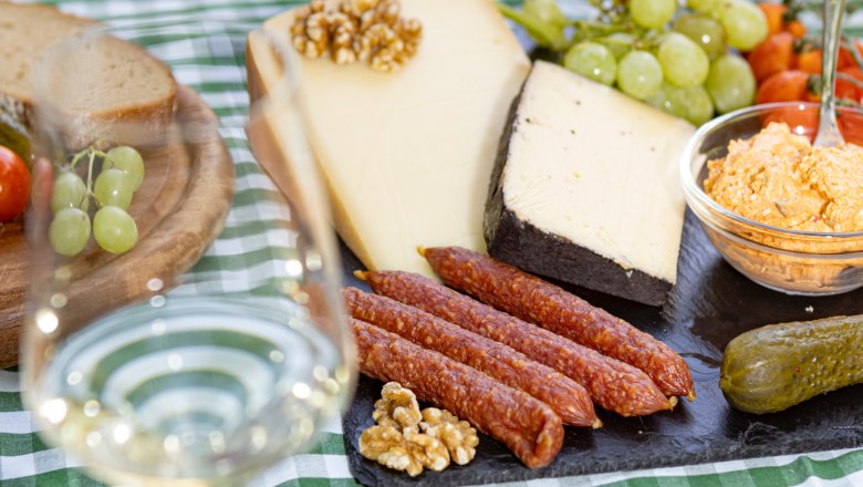 Heurigenjause, © Weinviertel Tourismus GmbH / Schwarz-König Picknicktisch mit Käse, Wurst, Brot, Weintrauben und einem Glas Weißwein auf kariertem Tischtuch.