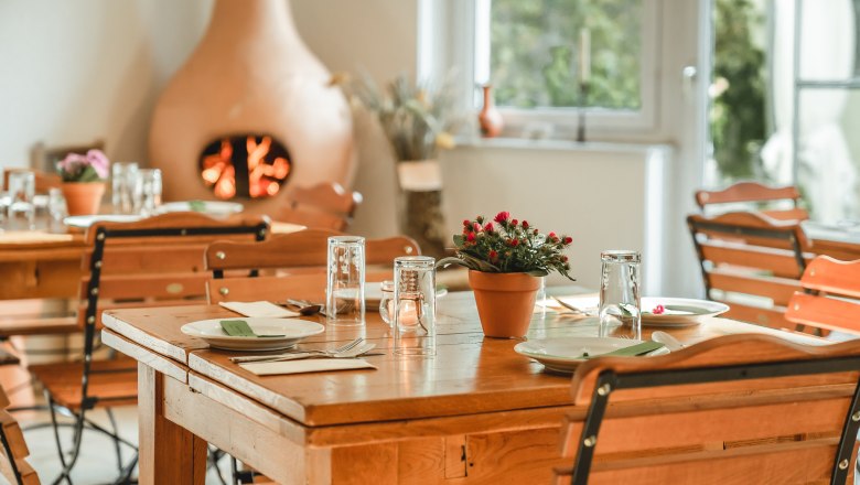 Breakfast room, © Matthias Auer A cozy breakfast room with wooden tables and chairs, set with crockery and glasses. A fireplace in the background creates a warm atmosphere.