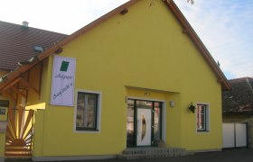 Außenaufnahme, © Fam Aigner Gelbes Gebäude mit einem Schild "Aigner Ausg'steckt is" an der Fassade.