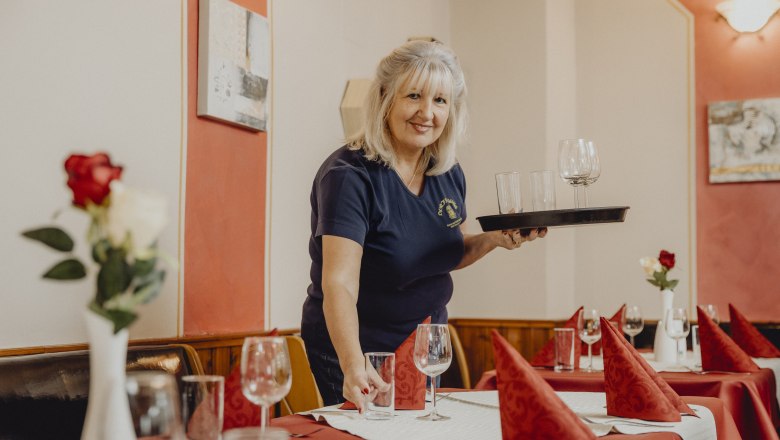 Landlady Erika Bischof, © Niederösterreich Werbung/Sophie Menegaldo Woman sets table in a restaurant with glasses and red napkins.