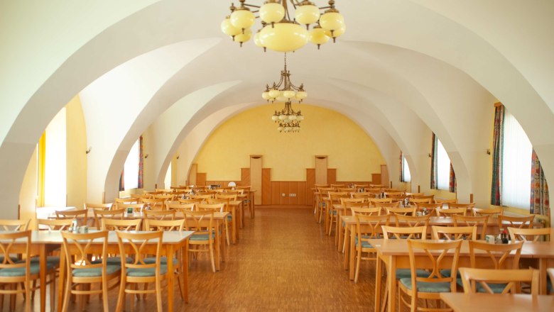 Gasthaus Bayer, © Clara Körner Innenansicht eines großen Speisesaals mit Holztischen und -stühlen, gelben Wänden und dekorativen Kronleuchtern.