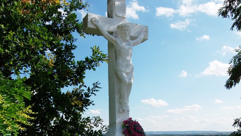 Zeißlerkreuz, © Weinsraße Weinviertel Weißes Steinkreuz mit Jesusfigur und Blumenkranz, umgeben von Bäumen und Landschaft im Hintergrund.