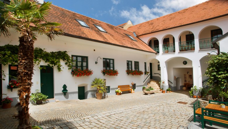 Gemütlicher Winzerhof, © Astrid Bartl Ein traditioneller österreichischer Innenhof mit weißen Gebäuden, roten Ziegeldächern und Blumen.