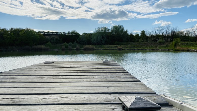 Idyllic fishing paradise, © Weinstraße Weinviertel Wooden footbridge at the brick kiln pond in Zellerndorf, surrounded by trees and a blue sky with clouds.