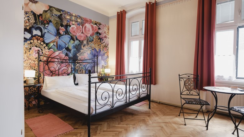 Oriental room, © Michael Reidinger A stylishly furnished room with a wrought-iron bed, floral wallpaper and a small table with chairs in front of large windows.