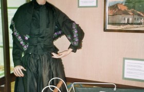 Trachtenpuppe, © Adolf Haider Eine Schaufensterpuppe in traditioneller Kleidung neben einem alten Kinderwagen in einem Ausstellungsraum.