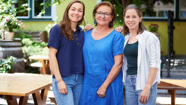 The Schmidt family, © Ben Leitner Three women stand smiling in a garden with wooden tables and plants.