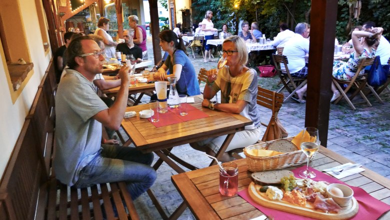 20ers Buschenschank, © Zwanziger Menschen sitzen an Holztischen in einem gemütlichen Außenbereich und genießen Essen und Getränke.