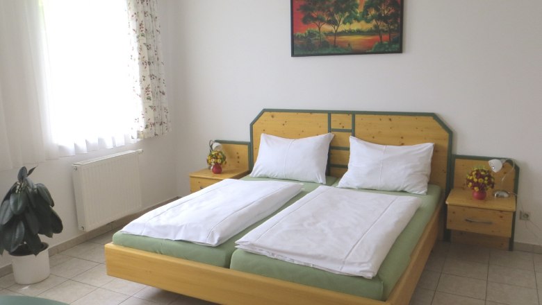 Gästezimmer Nexing, © Urani Ein Gästezimmer mit Doppelbett, Nachttischen, einem Bild an der Wand und einem Fenster mit Vorhängen.