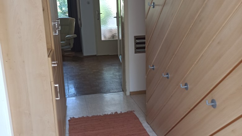 In the hallway, © Multimedia Lauda A hallway with wooden walls, hooks and a red carpet. A room with an armchair and a glass door is visible in the background.