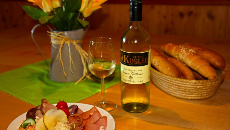 Heurigenjause, © Weingut Kugler Ein gedeckter Tisch mit Wein, Brot und einer Platte mit Aufschnitt.