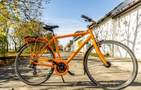 Radfahren im Weinviertel, © Weinviertel Tourismus GmbH / POV / Robert Herbst Ein orangefarbenes Fahrrad steht auf einer sonnigen Straße, umgeben von Bäumen und Gebäuden.