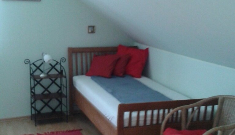 Dreibettzimmer (Einzelbett) bei Fam. Waysmayer, © © Bianca Waysmayer Ein Einzelbett in einem Zimmer mit Dachschräge, rotem Teppich, Rattanstuhl und Regal.