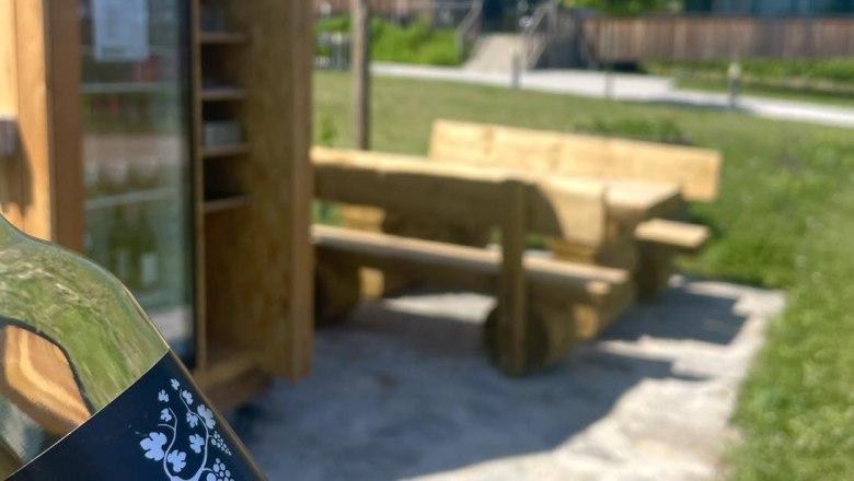 Cyclist's rest, © Weingut Grillmeier-Ladentrog A bottle labeled 'Spritzer rocks' in front of a rest area with wooden benches and an outdoor fridge.