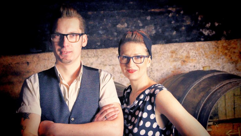 Pollak René und Ramona, © Matthias Karasek Ein Mann und eine Frau stehen nebeneinander vor einem rustikalen Hintergrund mit einem Fass.