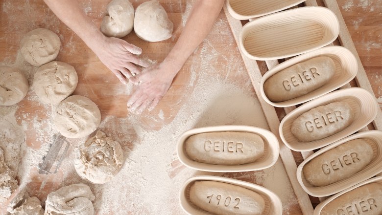 Echte Handarbeit, © Geier.die Bäckerei Hände formen Teig auf bemehltem Tisch, daneben Gärkörbe mit "GEIER" und "1902".