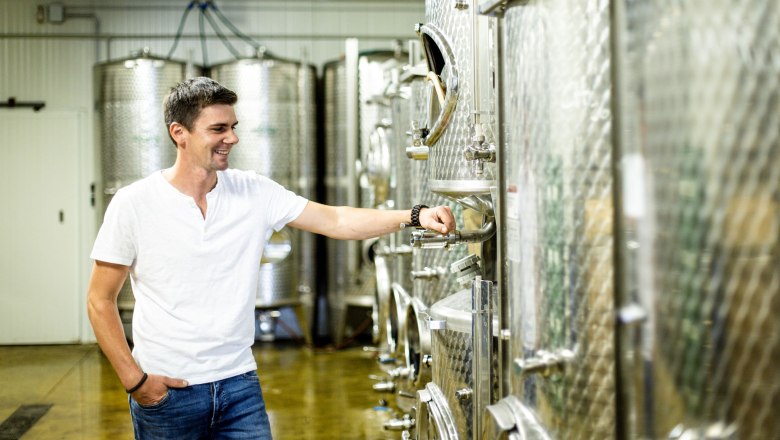 Winzer Roman Degn, © Astrid Bartl Ein Mann in einem Weinkeller steht lächelnd neben großen Edelstahltanks.
