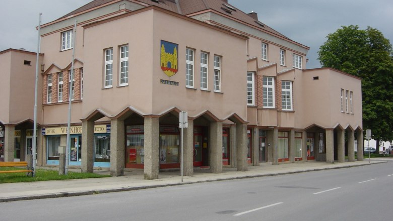 Rathaus, © Gemeinde Hohenau/ March Ein modernes Rathausgebäude mit rosa Fassade und großen Fenstern. Es hat ein Wappen an der Ecke und Geschäfte im Erdgeschoss.