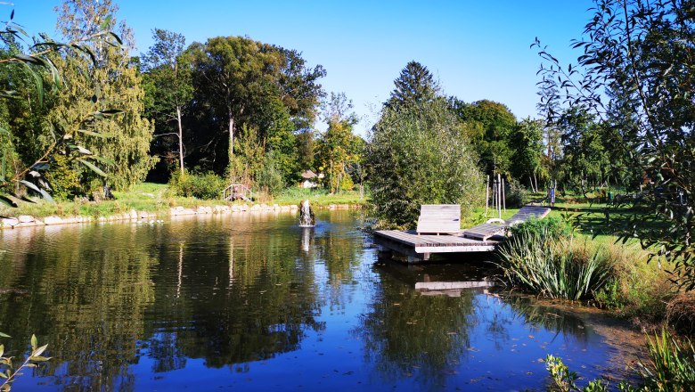Relaxing by the landscaped pond, © Weinstraße Weinviertel A tranquil pond with wooden loungers and lush vegetation under a blue sky.