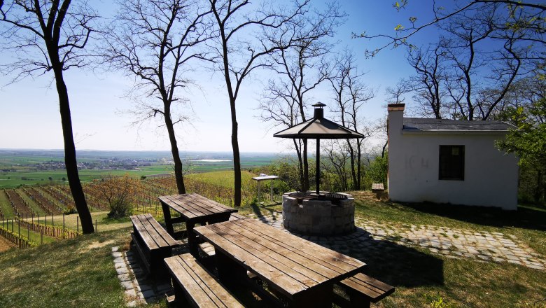 Verweilen am Hutberg, © Weinstraße Weinviertel Picknickplatz mit Holztischen und Grill auf einem Hügel mit Blick auf Weinberge und ein kleines Gebäude.