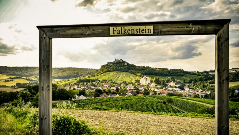 Blick auf Burgruine Falkenstein, © Weinviertel Tourismus / POV Robert Herbst Blick auf das Dorf Falkenstein mit Weinbergen und einer Burgruine im Hintergrund, eingerahmt von einem Holzrahmen mit der Aufschrift 'Falkenstein'.
