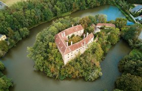 Burg Burgschleinitz, © Waldviertel Tourismus, lichtstark.com Luftaufnahme einer von Wasser umgebenen Burg mit roten Dächern und umgebenden Bäumen.