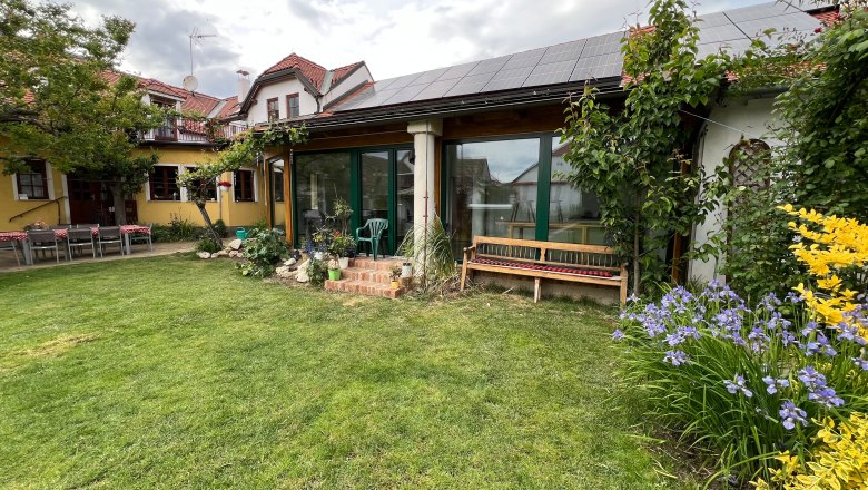 Garten, © Gästehaus Weinwurm Ein Garten mit Rasen, Blumenbeeten und einem Haus mit Solarpanelen auf dem Dach.