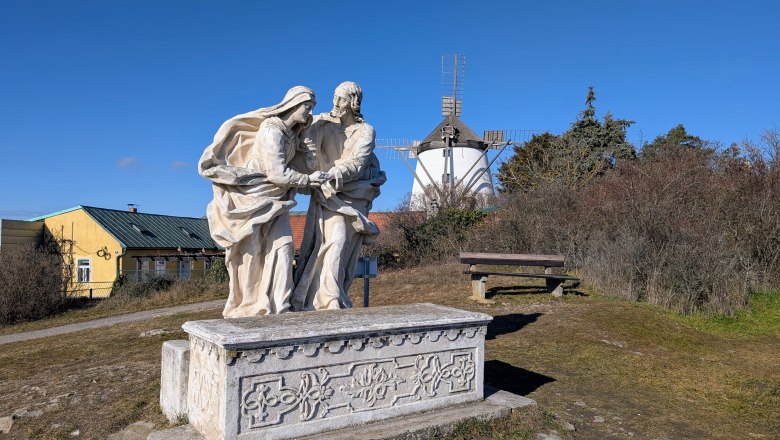 Kalvarienberg, © Retzer Land / Daniel Wöhrer Steinskulptur auf einem Hügel mit einer Windmühle im Hintergrund.