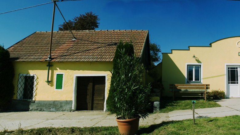 Weingut, © Rainer Schmid Zwei gelbe Gebäude eines Weinguts mit Ziegeldächern und Pflanzen im Vordergrund.
