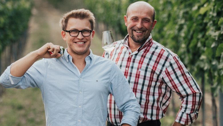 Winzer Paul und sein Vater sind ein starkes Team, © Winzerhof Zimmermann Zwei Männer stehen lächelnd in einem Weinberg, einer hält ein Weinglas.