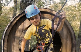 Kletterpark, © Fam. Freudhofmaier Ein Kind mit Helm und Kletterausrüstung in einem hölzernen Kletterpark-Tunnel.