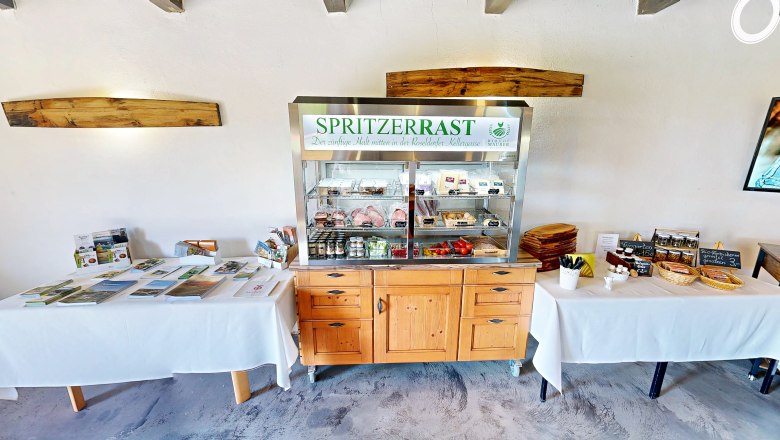 Spritzerrast – Campingstellplatz mit Jause und Panoramablick, © Bernhard Raab Ein Verkaufsstand mit regionalen Produkten und Informationsmaterialien in einem Raum mit Holzmöbeln.